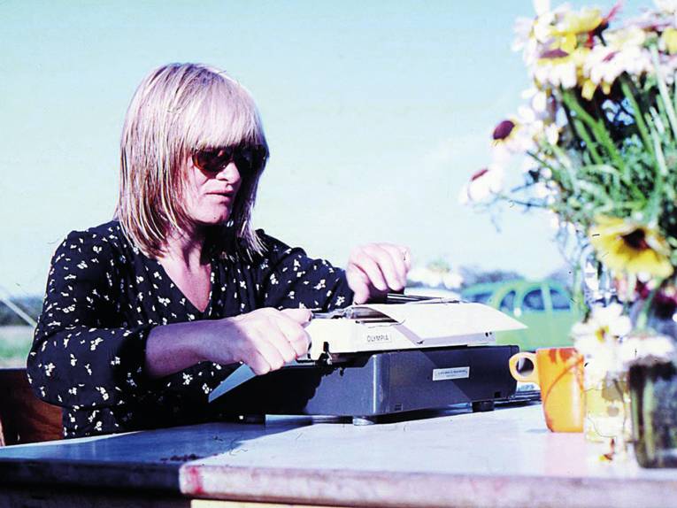 Zu sehen ist eine Person mit Pony und Sonnenbrille, die ein neues Blatt in eine Olympia Schreibmaschine einlegt.
